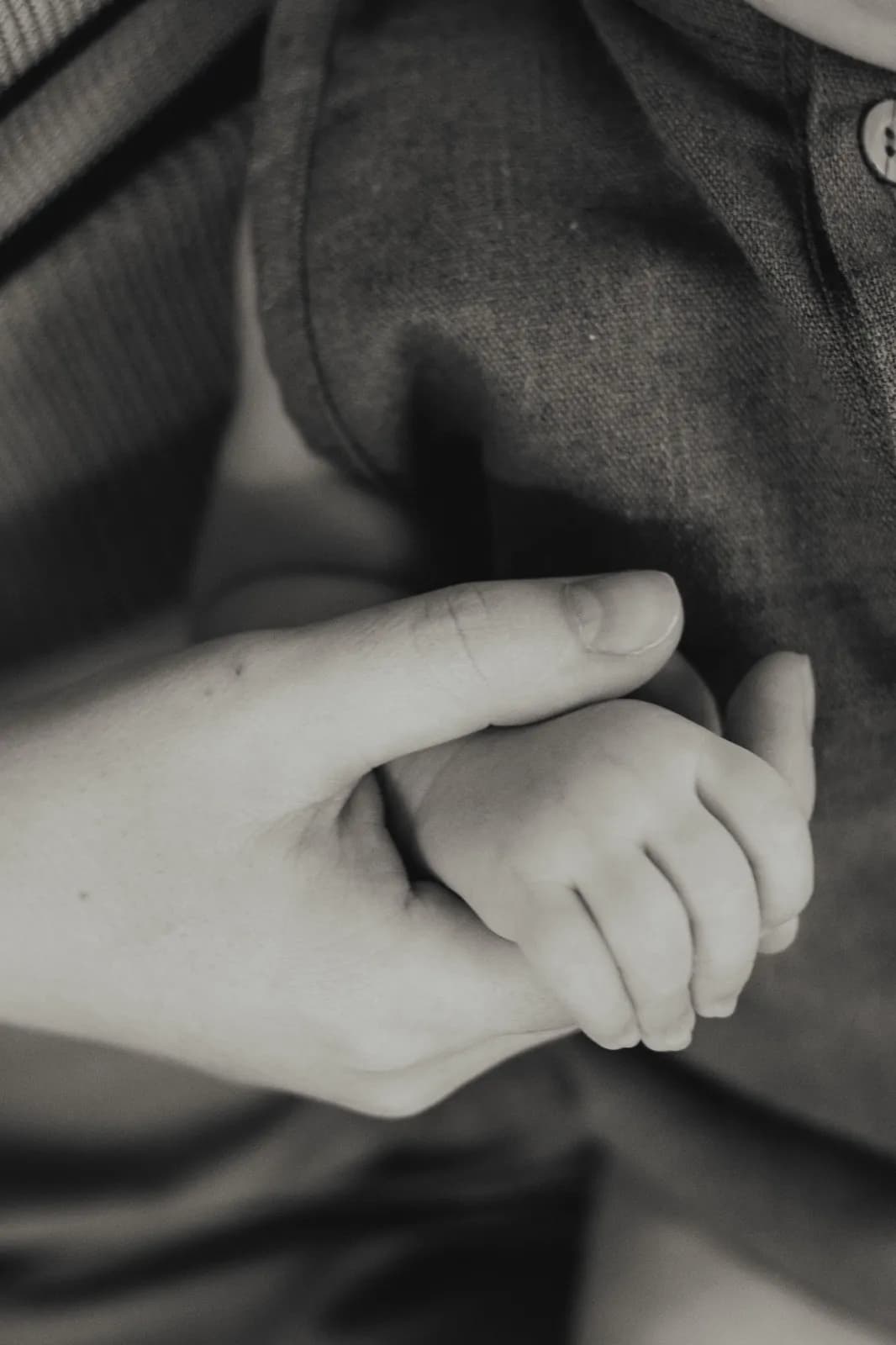 An adult hand gently holding a baby's hand against a linen shirt, in black and white